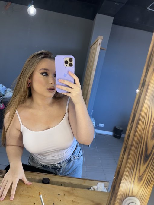A young woman takes a mirror selfie with a lavender iPhone, wearing a white tank top and jeans in what appears to be a dimly lit room.