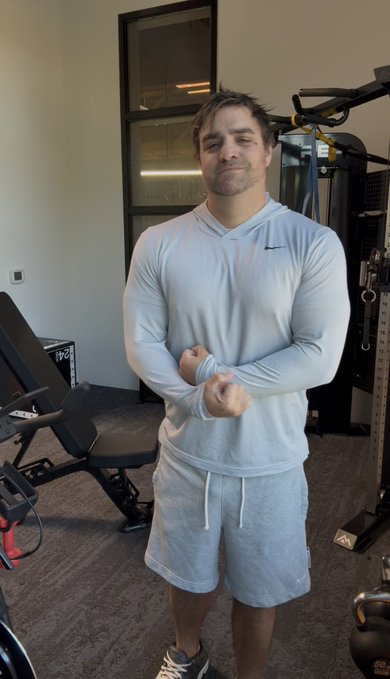 A man in a light blue long-sleeve Nike shirt and gray shorts stands in a gym, adjusting his sleeve.