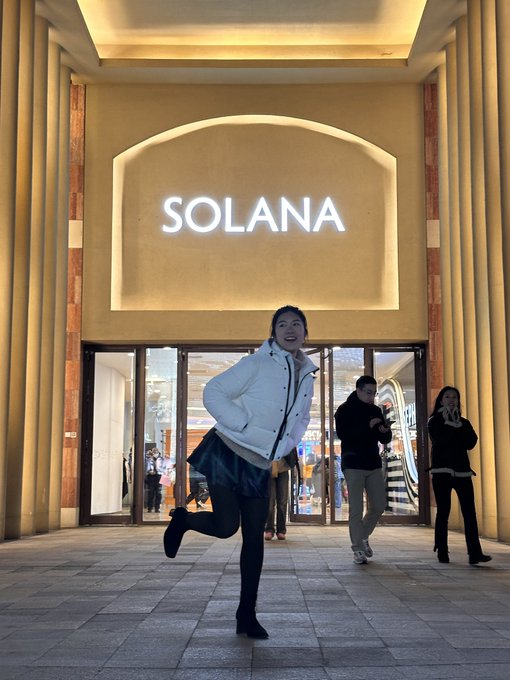 A person in a white puffer jacket and dark skirt poses playfully with one leg lifted in front of a building entrance displaying an illuminated 'SOLANA' sign.