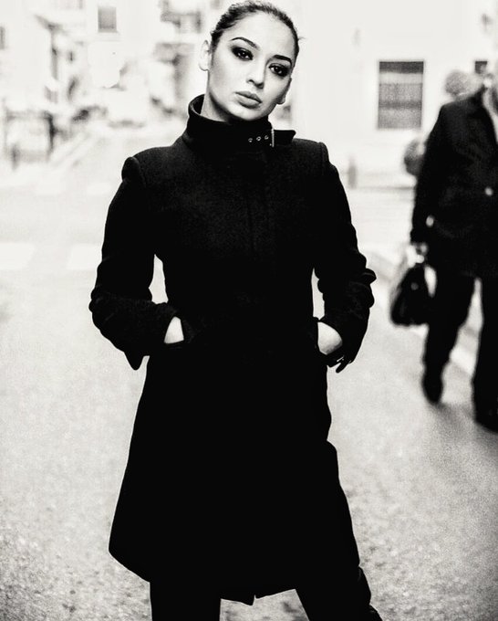 A young woman stands on a city street in a long dark coat, her hair pulled back and a serious expression on her face, while a blurred figure walks behind her. The somber black-and-white composition and her focused gaze evoke the bold, no-nonsense style associated with GN.