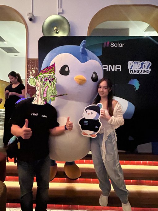 Two people posing with thumbs up in front of a large Pudgy Penguins mascot; one person's face is obscured by a cartoon graphic, and they are holding a small penguin cutout.