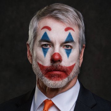 A portrait of a middle-aged man with gray hair and a beard wearing clown makeup with blue diamond shapes over the eyes and a red nose and mouth, dressed in a suit with an orange tie.