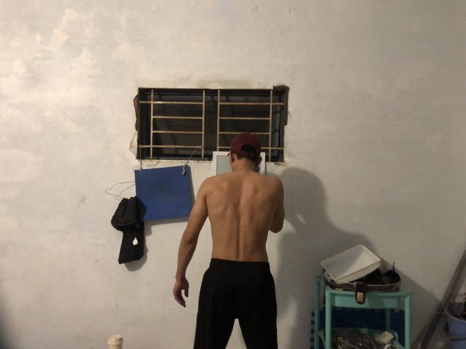 A shirtless man wearing a red cap backwards and black pants stands facing a wall with a barred window and hanging items.