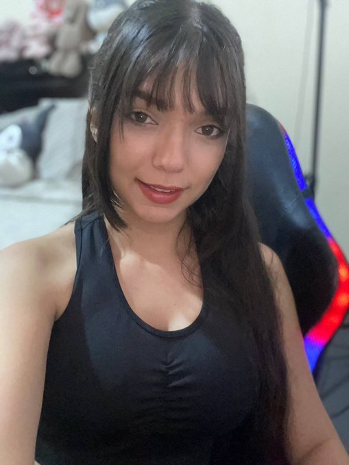 A young woman with long dark hair wearing a black tank top smiles softly at the camera in what appears to be a gaming setup, with a plush toy visible in the background. The image pairs with a cheerful morning greeting to Bitcoin holders, suggesting optimism about BTC's performance and a lighthearted jab at those who sold or attempted to trade against the trend.