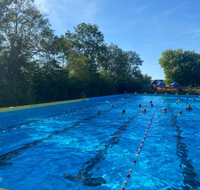 A large outdoor swimming pool marked with lane dividers where several people are swimming and participating in water activities, surrounded by trees under a clear blue sky.