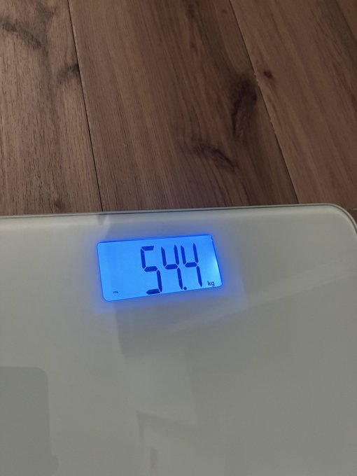 A digital bathroom scale displays a weight reading of 54.4 kg on its blue-lit LCD screen, resting on a wooden floor.