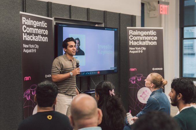 A speaker presents at the 'Raingentic Commerce Hackathon' event in New York City on August 8-9, with banners showing sponsor logos and an 'Investor Keynote' displayed on the screen behind him.