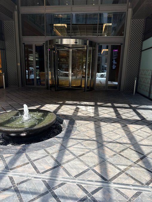 A modern building entrance features a revolving glass door with a small water fountain in the foreground, set against a patterned stone plaza. This image, shared as a "GM" (good morning) post, showcases an elegant architectural space that conveys a polished, professional start to the day.