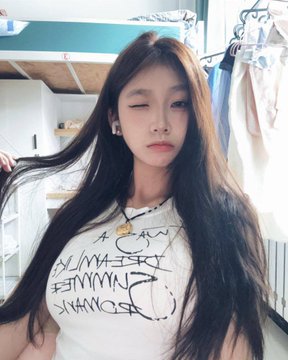 A selfie of a young woman with long dark hair, eyes closed, wearing a white t-shirt with black handwritten-style text and a graphic print, in what appears to be a room with hanging clothes.
