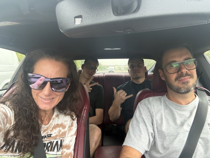 A casual selfie taken inside a car shows four people smiling and gesturing at the camera, with two adults in the front seats (a woman in sunglasses and a bearded man with glasses) and two young men in the back.