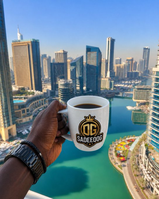A hand holds a white mug with a gold 'QC SADEEQOG' logo, filled with black coffee, overlooking a city skyline and marina.