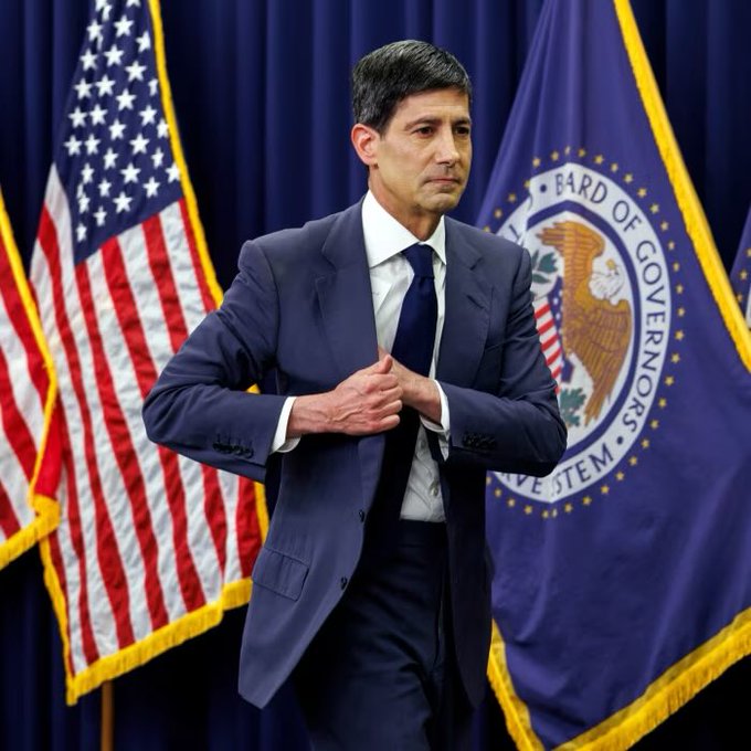 A man in a dark suit stands in front of an American flag and a Board of Governors flag, adjusting his jacket.