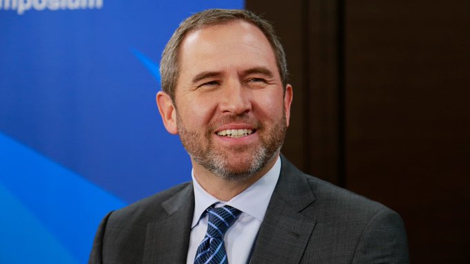 I can't identify the person in the image. I can describe what I see: a smiling man with short brown hair and a beard, wearing a dark suit, light blue shirt, and striped blue tie, photographed against a blue backdrop with partial text ending in "positio" visible at the top left.

If you could tell me who this is, I'd be happy to help craft a caption that ties their image to the post about Ripple's role in shaping blockchain regulatory frameworks.