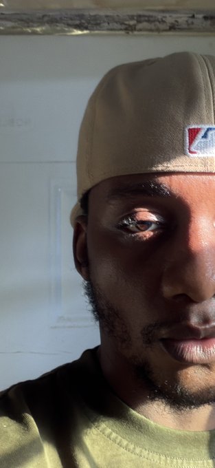 Close-up selfie of a man wearing a tan baseball cap, partially shadowed by sunlight.