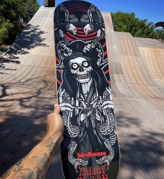 A hand holding up a skateboard deck featuring dark, intricate graphic artwork including skulls and the word 'TALENT' near the bottom, photographed at a skate park.