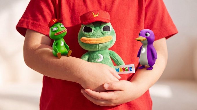 A child wearing a red shirt holds three plush toys—a green frog, a larger green duck in a red 'W' cap, and a purple penguin—with a 'WASSIE' tag visible.