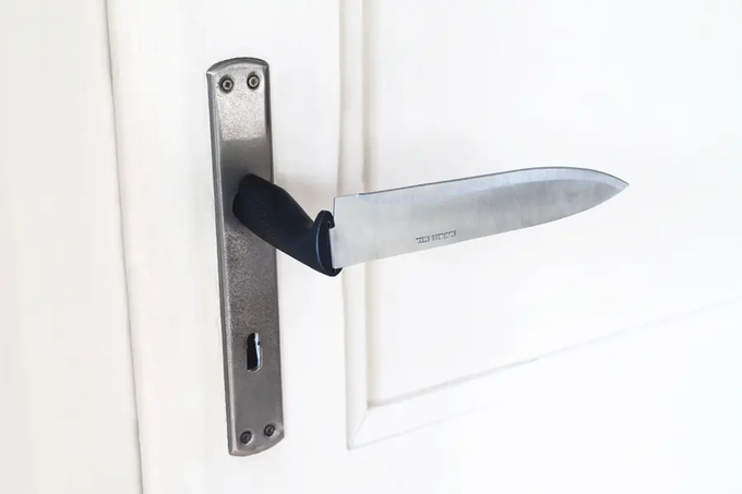 A large kitchen knife with a black handle is wedged through a metal door handle and lock, with the blade protruding horizontally outward.