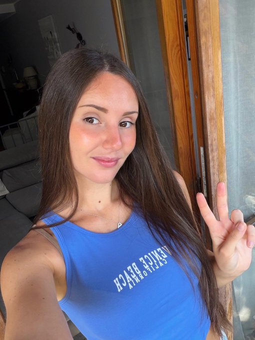 A selfie of a woman with long brown hair wearing a blue tank top that reads "AERIE BEACH CALIFORNIA" (partially reversed) and a silver heart necklace, making a peace sign with her hand.