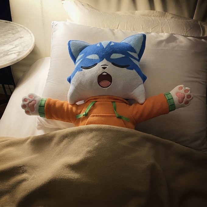 A plush toy of a sleeping blue and white cat character wearing an orange hoodie lies in bed with its arms outstretched, covered by a beige blanket.