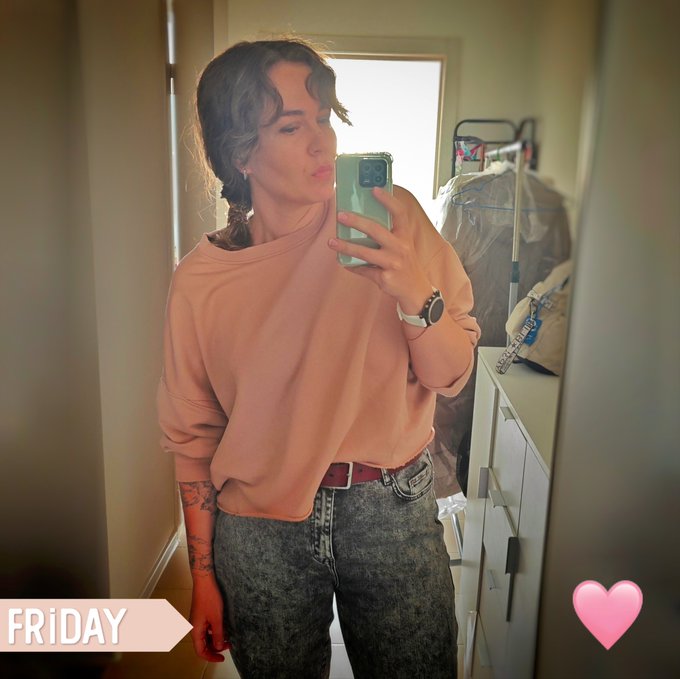 A mirror selfie of a woman in a peach sweater and grey jeans, labeled 'FRIDAY' with a pink heart emoji.