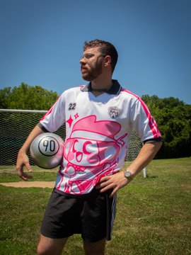 A bearded man wearing a vibrant pink and white soccer jersey featuring a stylized "CS" logo, the number 22, and a Chimpers design stands on a grassy field holding a soccer ball marked with "90." The bold jersey showcases the limited-edition drop from Chimpers Jerseys, whose striking design reflects the exclusive first-come, first-served release available through @dyli_io.
