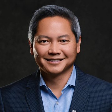 A professional headshot of a smiling man with graying hair wearing a blue shirt and dark blazer.