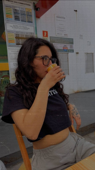 A young woman with glasses and long dark hair, wearing a black crop top and grey shorts, drinking from a paper cup while sitting in front of a wall with informational notices.