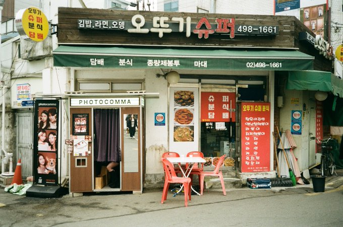 A Korean storefront with signage including '오뚜기 슈퍼' and 'PHOTOCOMMA', showing a photo booth with sample portraits, red plastic chairs, and a menu display.