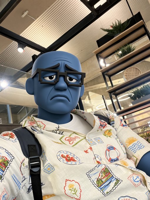 A 3D animated blue-skinned character with thick square glasses and a sad expression wears a patterned white shirt.