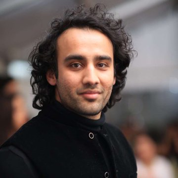 A close-up portrait of a man with curly dark hair wearing a black collared shirt.