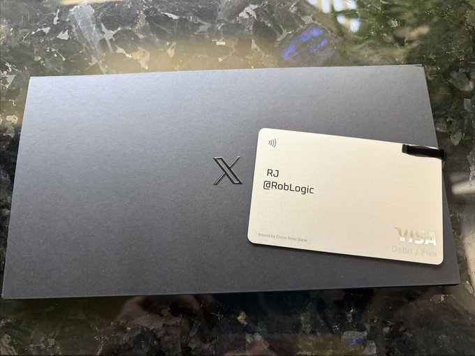 A sleek dark gray box with the X logo displayed next to a metallic Visa debit card labeled 'RJ @RobLogic' on a marble surface.