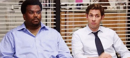 Two men, dressed in blue and white, sit at a desk with closed blinds, engaged in conversation or observing something outside the window.