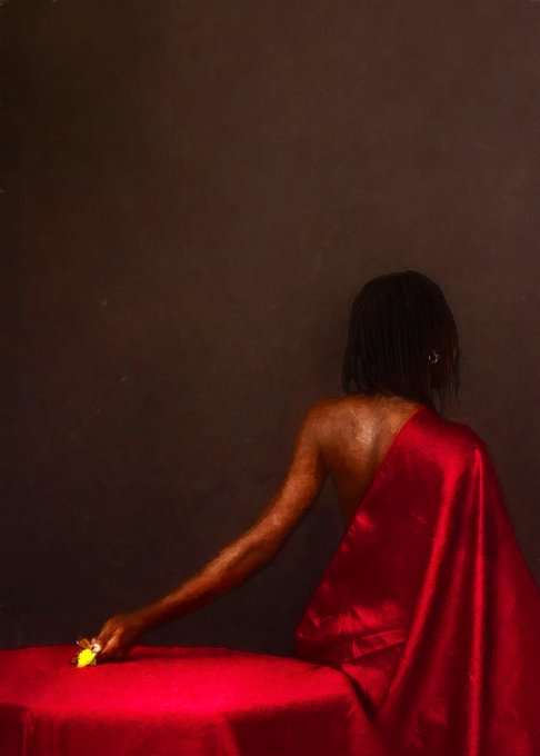A person with dark hair in a red satin dress sits at a red table, their face turned away, reaching toward a small yellow flower in dramatic lighting against a dark background.