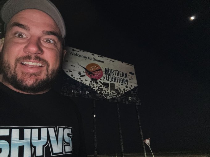 A man in a beanie and SHYVS t-shirt smiles excitedly in front of a 'Welcome to the Northern Territory' sign at night.