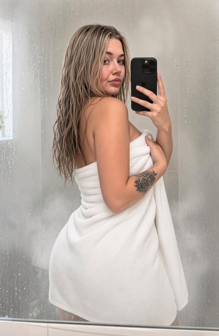 A mirror selfie of a woman with wet blonde hair wrapped in a white towel, revealing a tattoo on her upper arm.