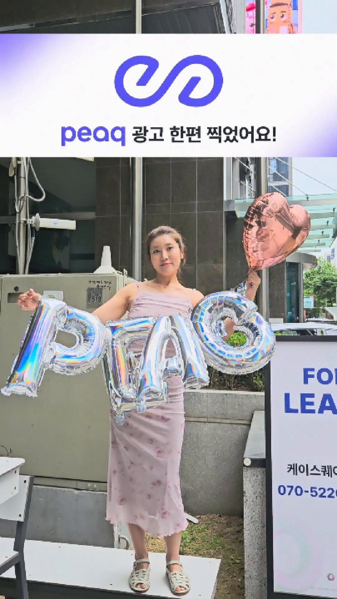 In this image, there's a woman standing outside holding balloons shaped like letters "P" and "A", which are made of foil material. She appears to be posing for a photo with these unique balloons. The background features a building, suggesting that she is in an urban setting or possibly at an event taking place inside the building. A sign can also be seen behind her, although its contents are not visible in this particular image.