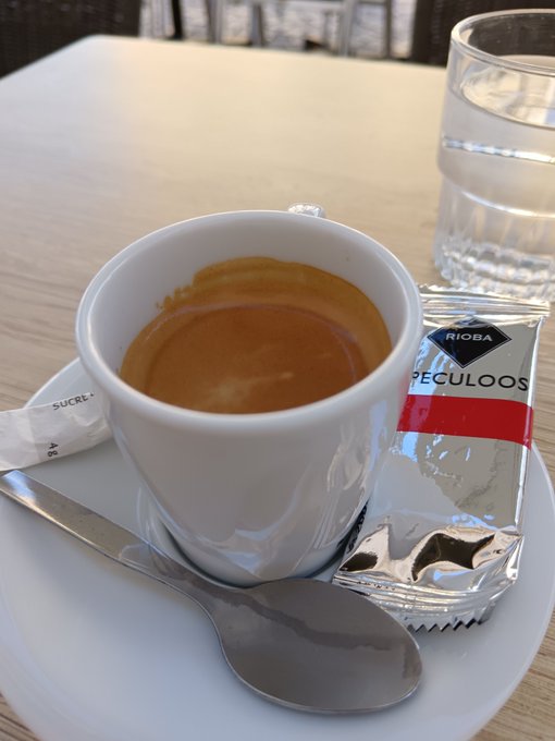 A small white cup of espresso on a saucer with a spoon, accompanied by a glass of water and a 'SPECULOOS' syrup packet from RIOBA, likely at an airport lounge.