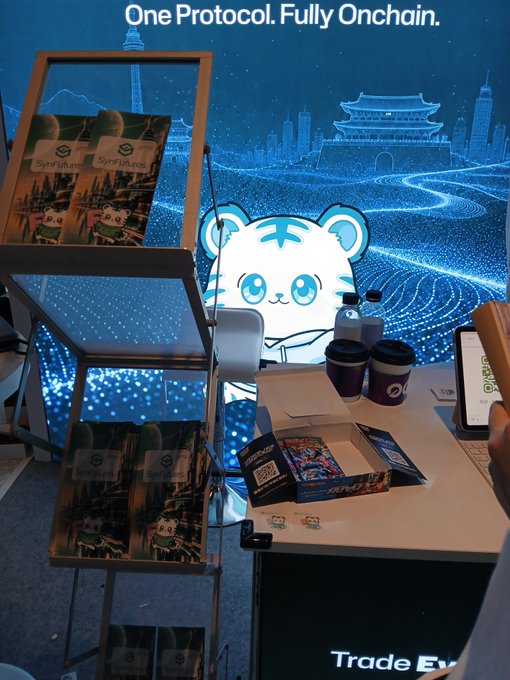 A trade show booth display for SynFutures with the tagline 'One Protocol. Fully Onchain,' featuring a cute tiger mascot character, promotional materials on shelves, and a digital cityscape backdrop.
