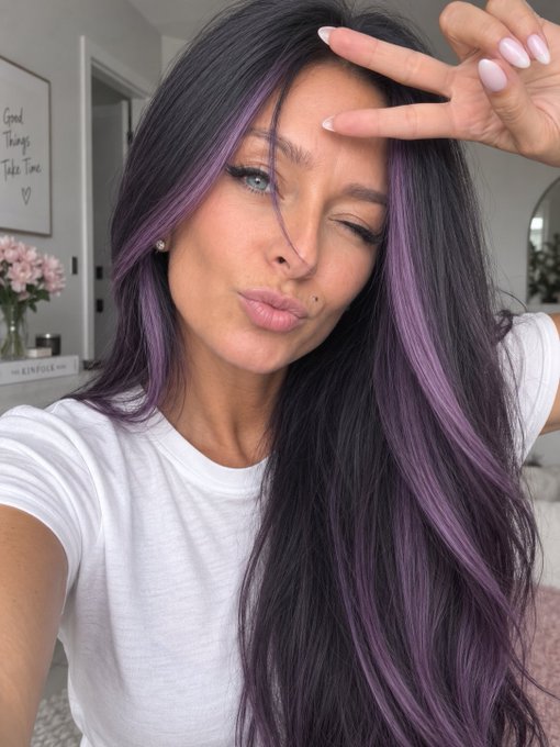 A close-up selfie of a woman with long dark hair featuring purple highlights, making a peace sign over one eye and puckering her lips, with a 'Good Things Take Time' sign in the background.