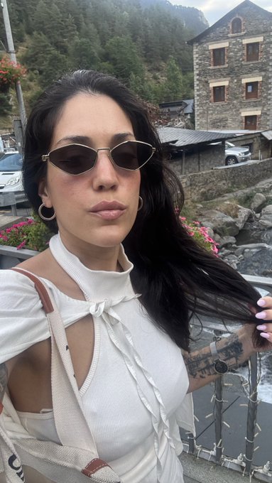 Selfie of a woman with long dark hair wearing sunglasses and a white top with bow detailing, standing near a stone building with mountains in the background.