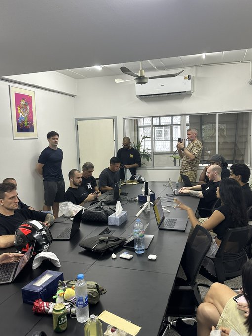A group of people gather around a long conference table in a meeting room with laptops, water bottles, and bags scattered about, while one person stands filming with a phone.