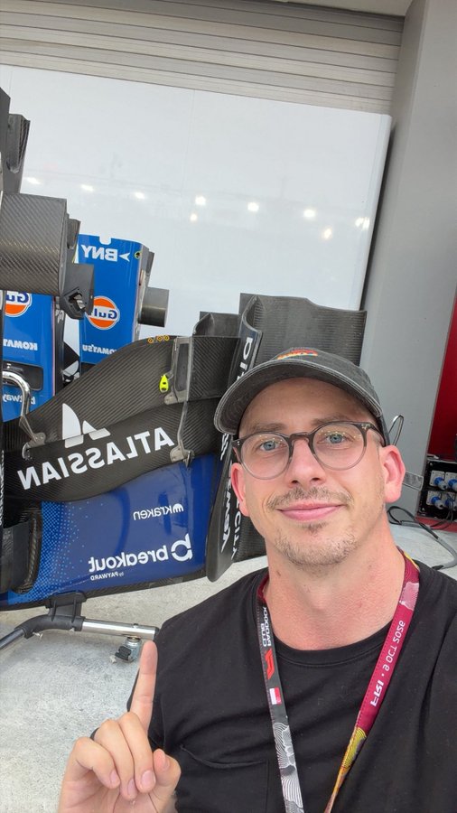 A man in a cap and glasses smiles and gives a thumbs-up in a racing team's garage, surrounded by BWT Alpine F1 car parts with Gulf and Castrol sponsorship logos visible, capturing the excitement of being behind the scenes in the pit lane.