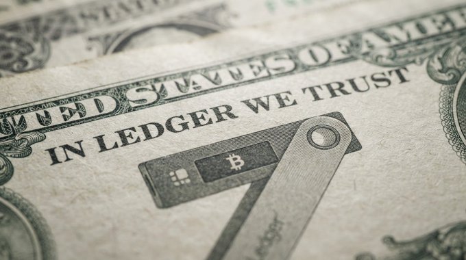 A close-up of a US dollar bill with the phrase 'IN LEDGER WE TRUST' replacing the standard text, alongside an illustration of a hardware cryptocurrency wallet.