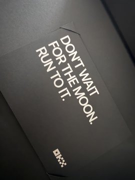 A sleek black OKX-branded card or box featuring the motivational text "DON'T WAIT FOR THE MOON, RUN TO IT." alongside the OKX logo, symbolizing the crypto exchange's brand ethos of proactive ambition. The image relates to the post as a piece of OKX merchandise that the author wishes to receive again, expressing envy toward "Lao Zhang" for getting OKX swag while feeling sidelined by the exchange.