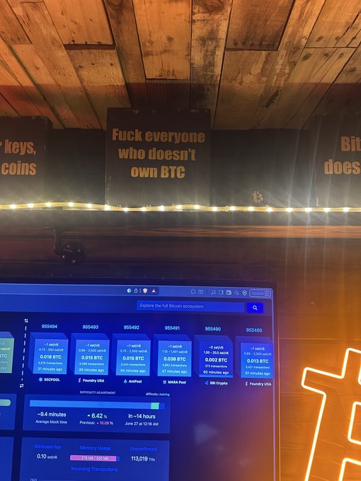 A monitor displays a Bitcoin block explorer dashboard showing blocks 955494–955489 with a difficulty rating of ~9.4 minutes and mempool fees around 10-14 sats/vB, sitting below a defiant sign reading "Fuck everyone who doesn't own BTC"—fittingly illustrating the poster's rejection of fiat currency and full embrace of Bitcoin since 2013.