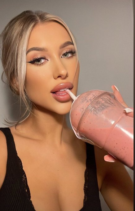 A portrait of a blonde woman with dramatic makeup sipping a beverage from a pink protein shaker bottle.