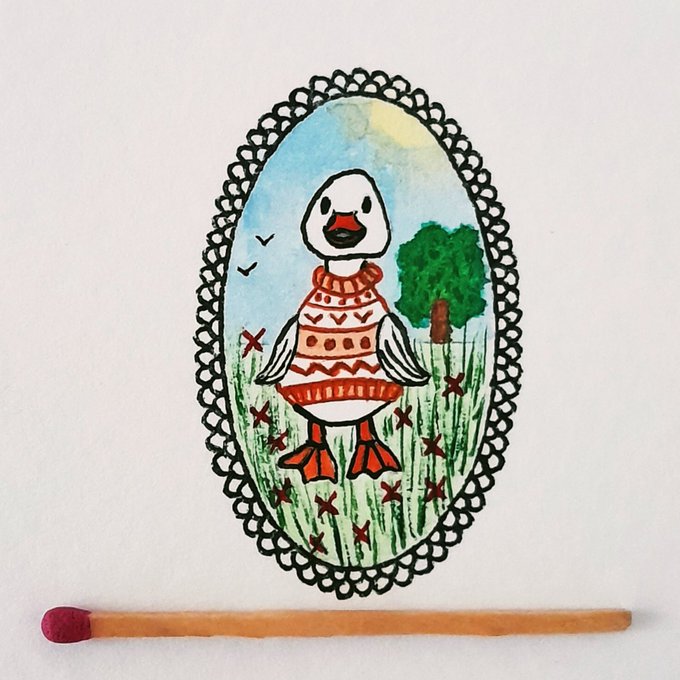 A hand-drawn illustration of a duck wearing an orange and red patterned sweater, framed in an ornate oval border with a tree and grass scene, with a wooden matchstick placed below for scale.