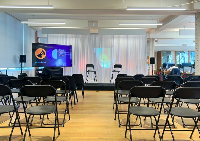 An empty event venue with rows of black folding chairs facing a small stage with a TV screen displaying "Official Corporate Sponsor" and "wewhost" logos, set up in a bright industrial-style room.
