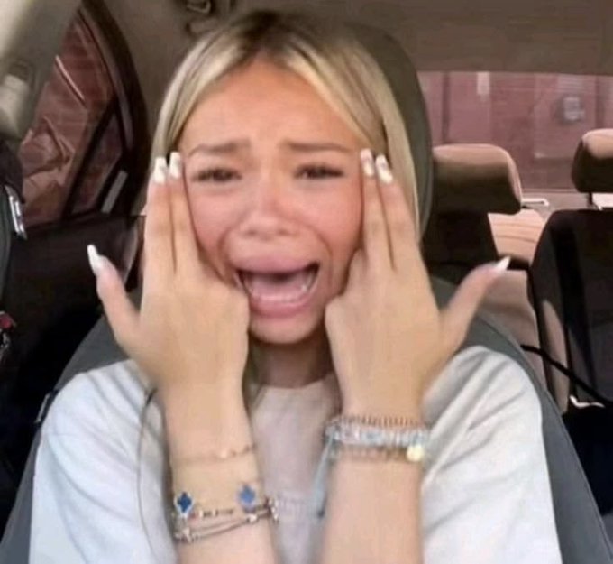 A young woman with blonde hair sits in a car, holding her face with both hands in a dramatic crying or screaming expression, wearing bracelets and a light-colored top.