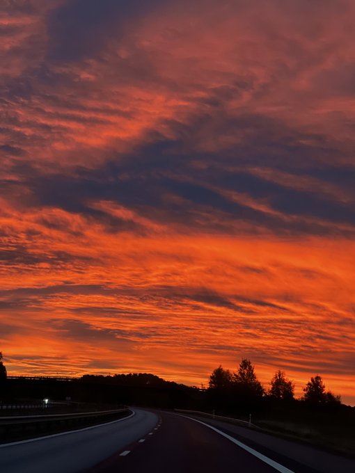 A vibrant orange and red sunset sky filled with clouds above a curving highway and silhouetted trees.